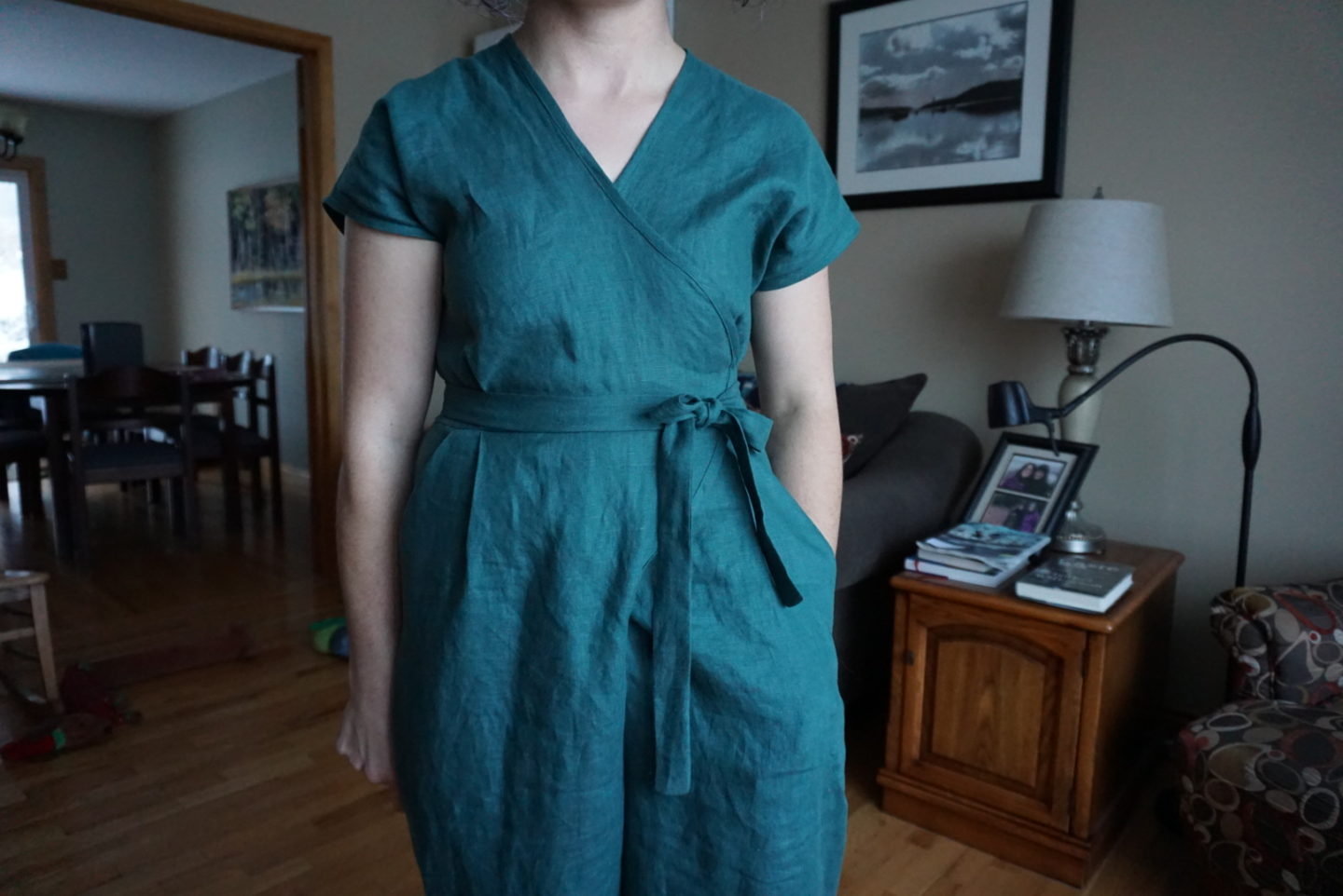 Megan is wearing a forest green wrap jumpsuit, standing in front of a couch in a living room.