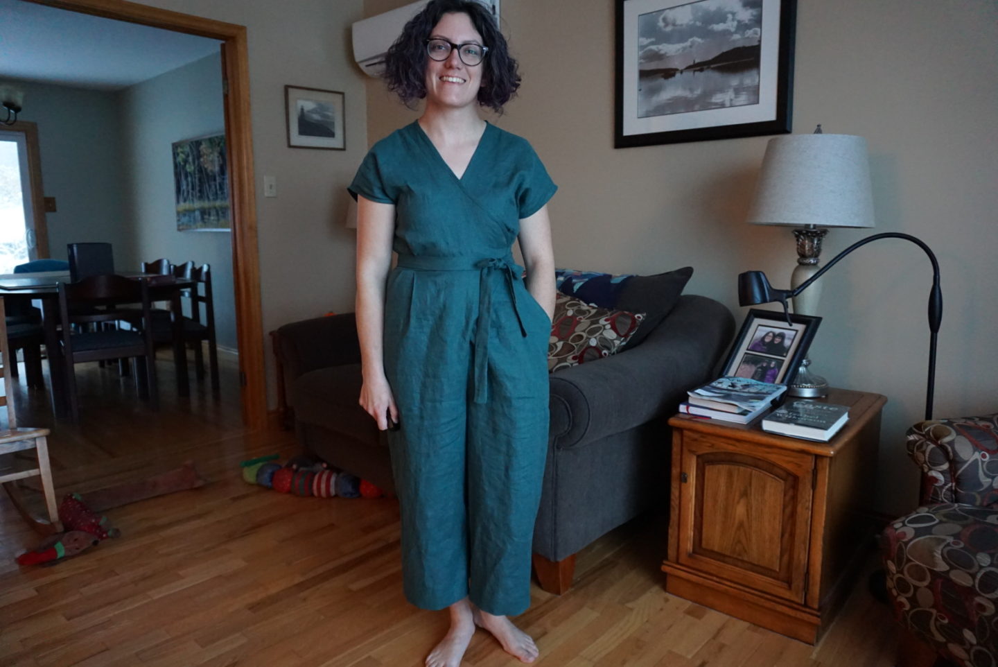 Megan stands in front of a couch in her living room. She is wearing a forest green wrap jumpsuit.